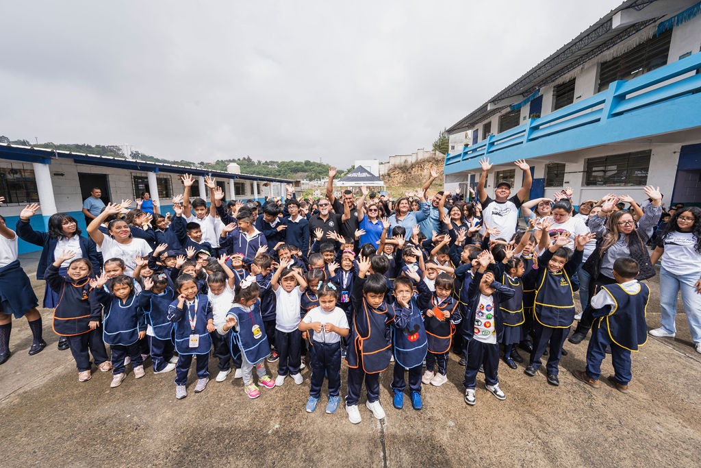 A large group of enthusiastic children and adults wave joyfully outside a school, celebrating together on a cloudy day.
