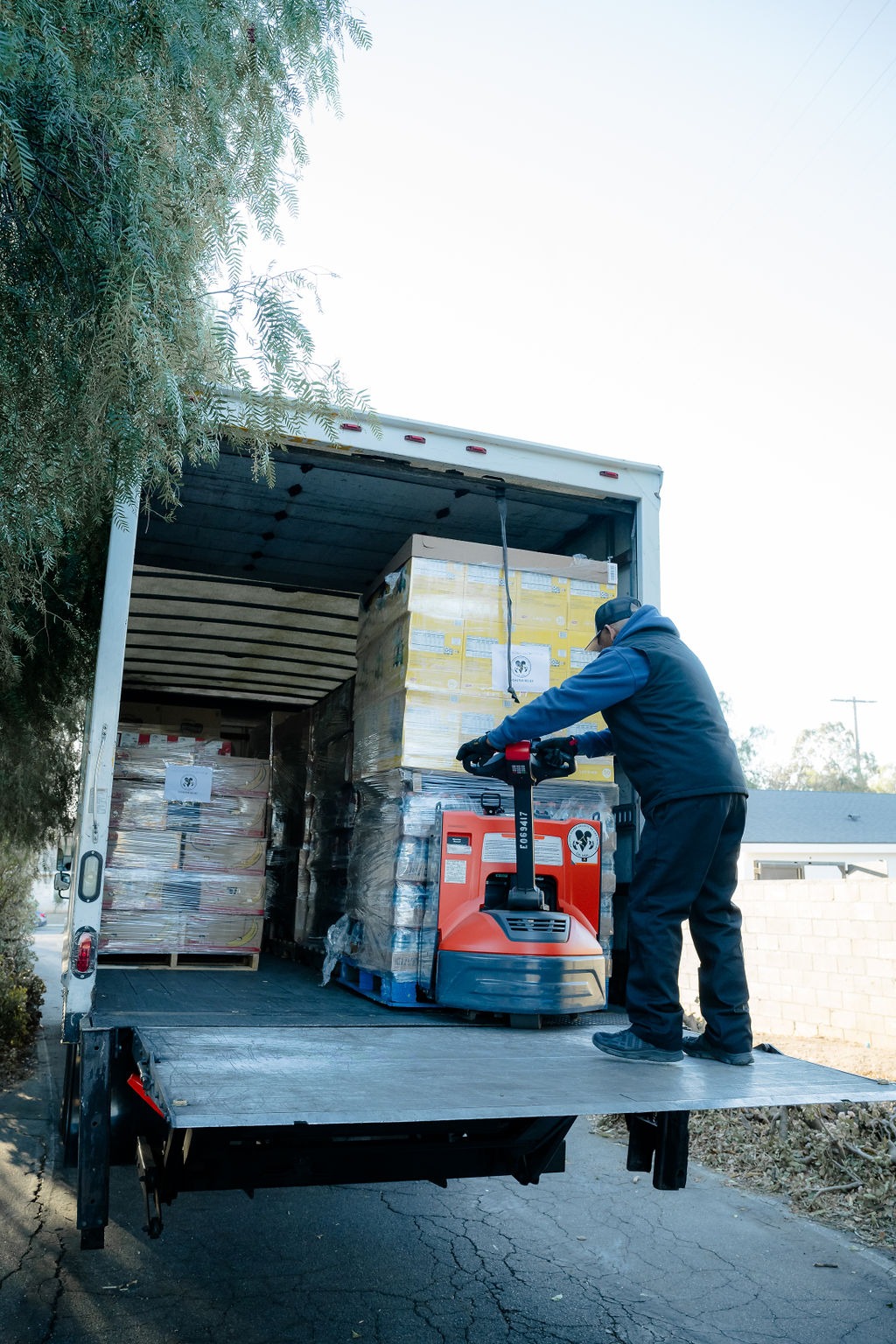 A worker operates a pallet jack to unload stacked boxes from a delivery truck, showcasing logistics and transportation operations.