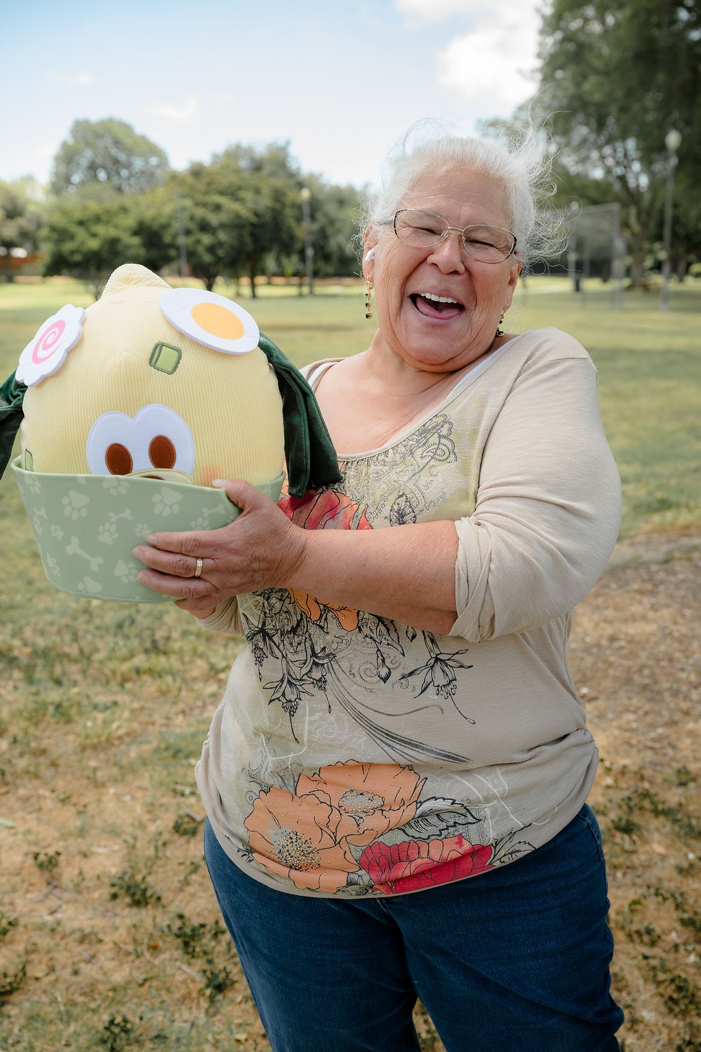 A smiling older woman holds a colorful, plush toy in a park, showcasing joy and a sense of playfulness, highlighting community engagement.