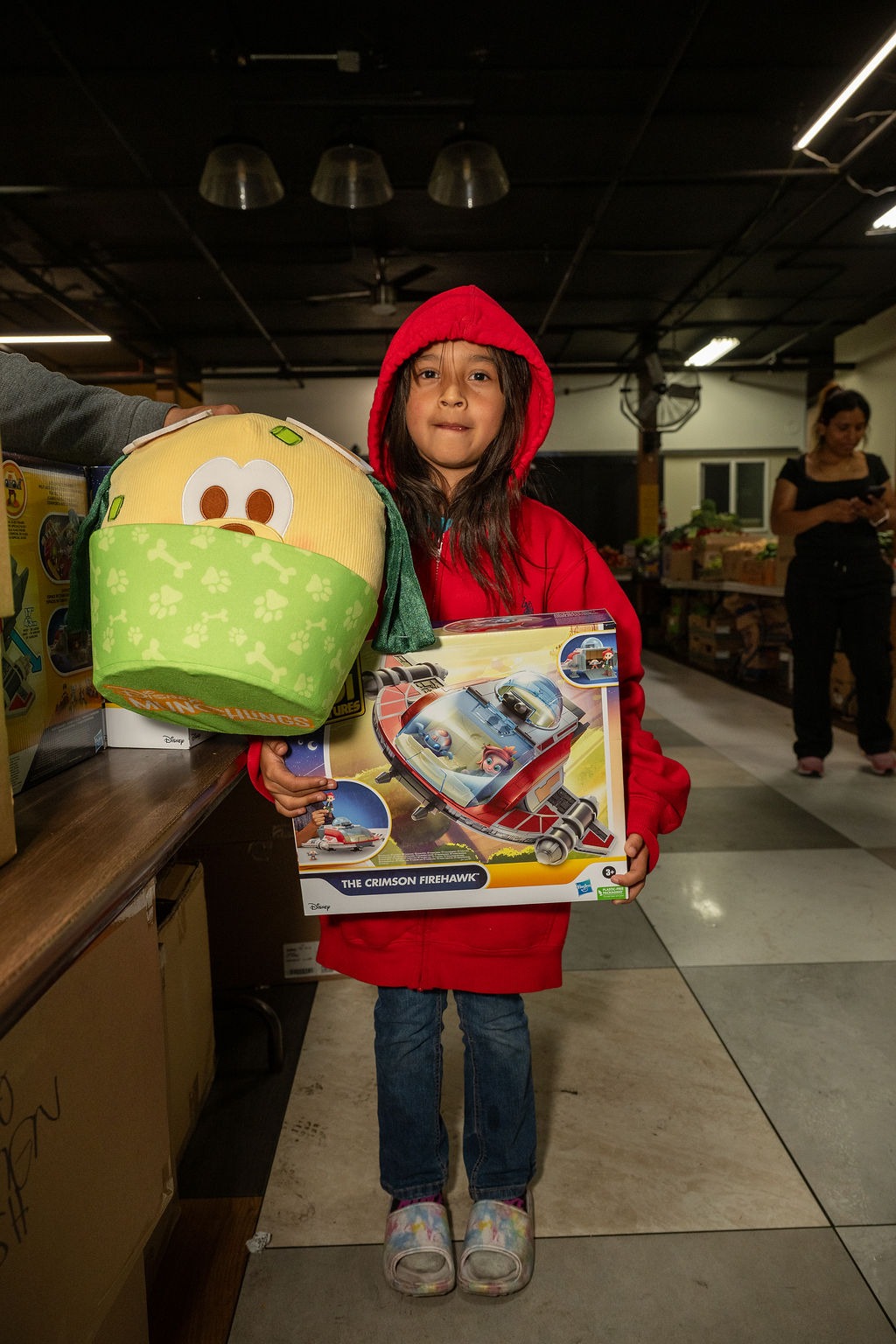 A girl in a red hooded jacket holds a toy box labeled "The Crimson Firehawk" and a plush toy, smiling in a store filled with products.