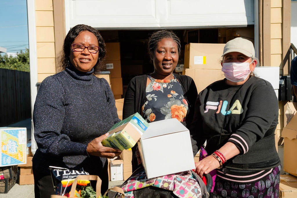 Three women smile while holding food and a package in front of a garage filled with boxes, highlighting community support efforts.