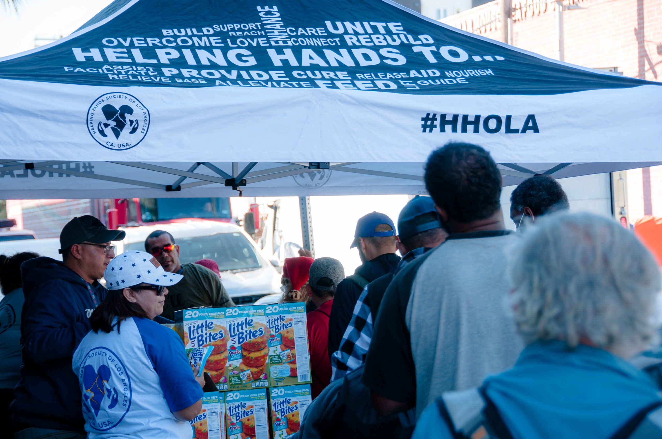 People line up under a tent with the Helping Hands logo, receiving food packages from the Helping Hands Society of Los Angeles.
