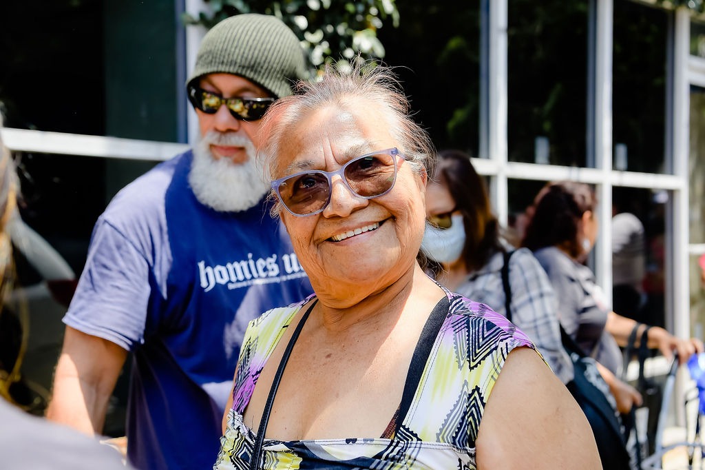 Smiling elderly woman wearing colorful attire and glasses, standing outdoors with a diverse group, emphasizing community connection and warmth.