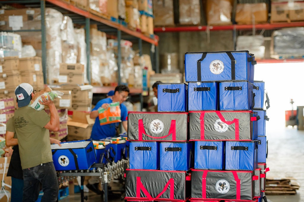Volunteers organize blue and black insulated bags filled with supplies in a warehouse, facilitating food distribution efforts.