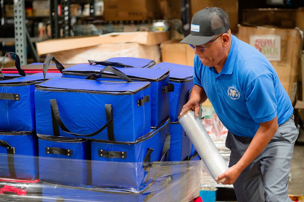 A warehouse worker is lifting a silver container while handling stacks of blue insulated bags, preparing for distribution or delivery tasks.