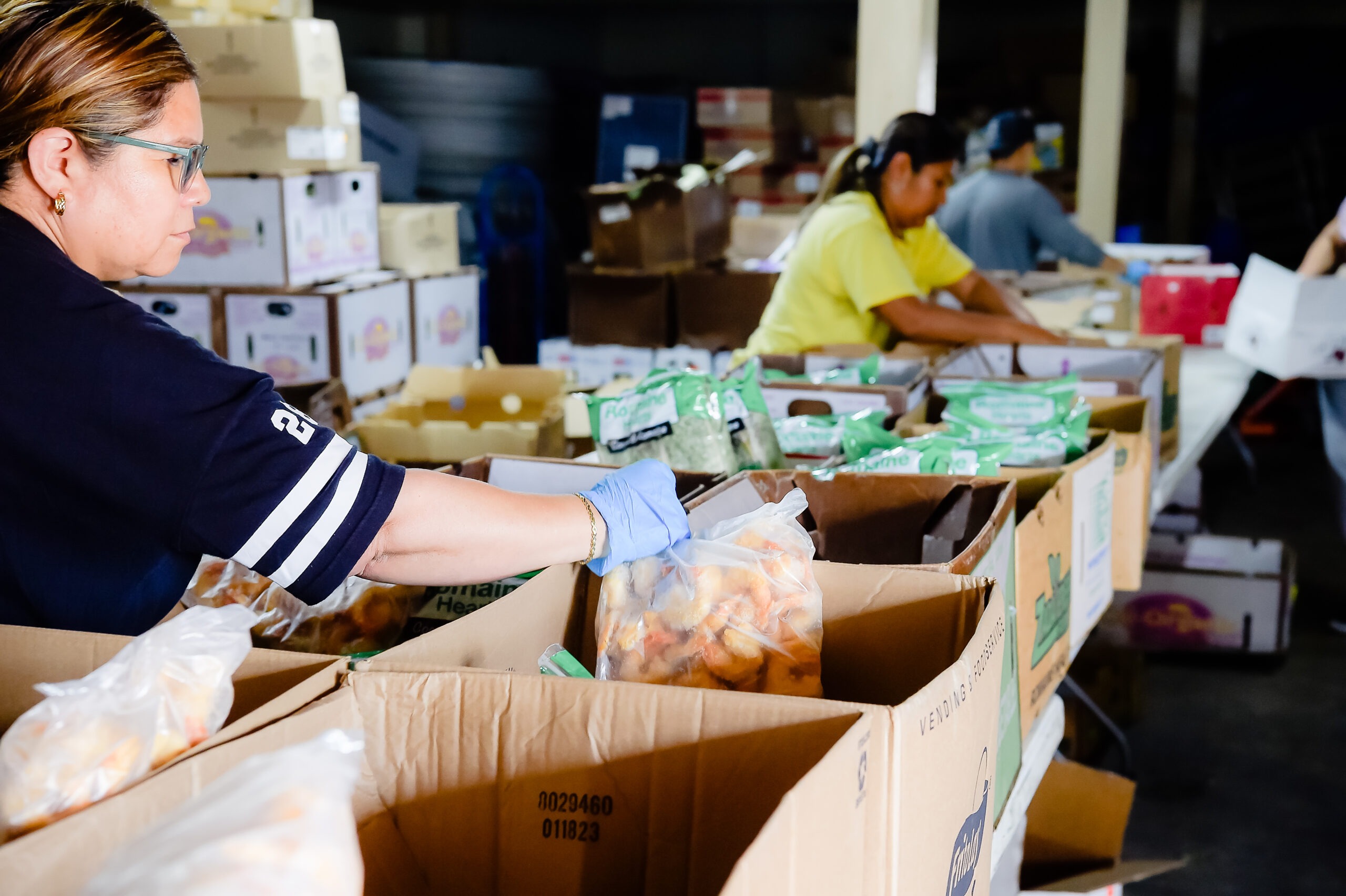 Volunteers sort and package food items in a warehouse, highlighting efforts in food distribution and community support.