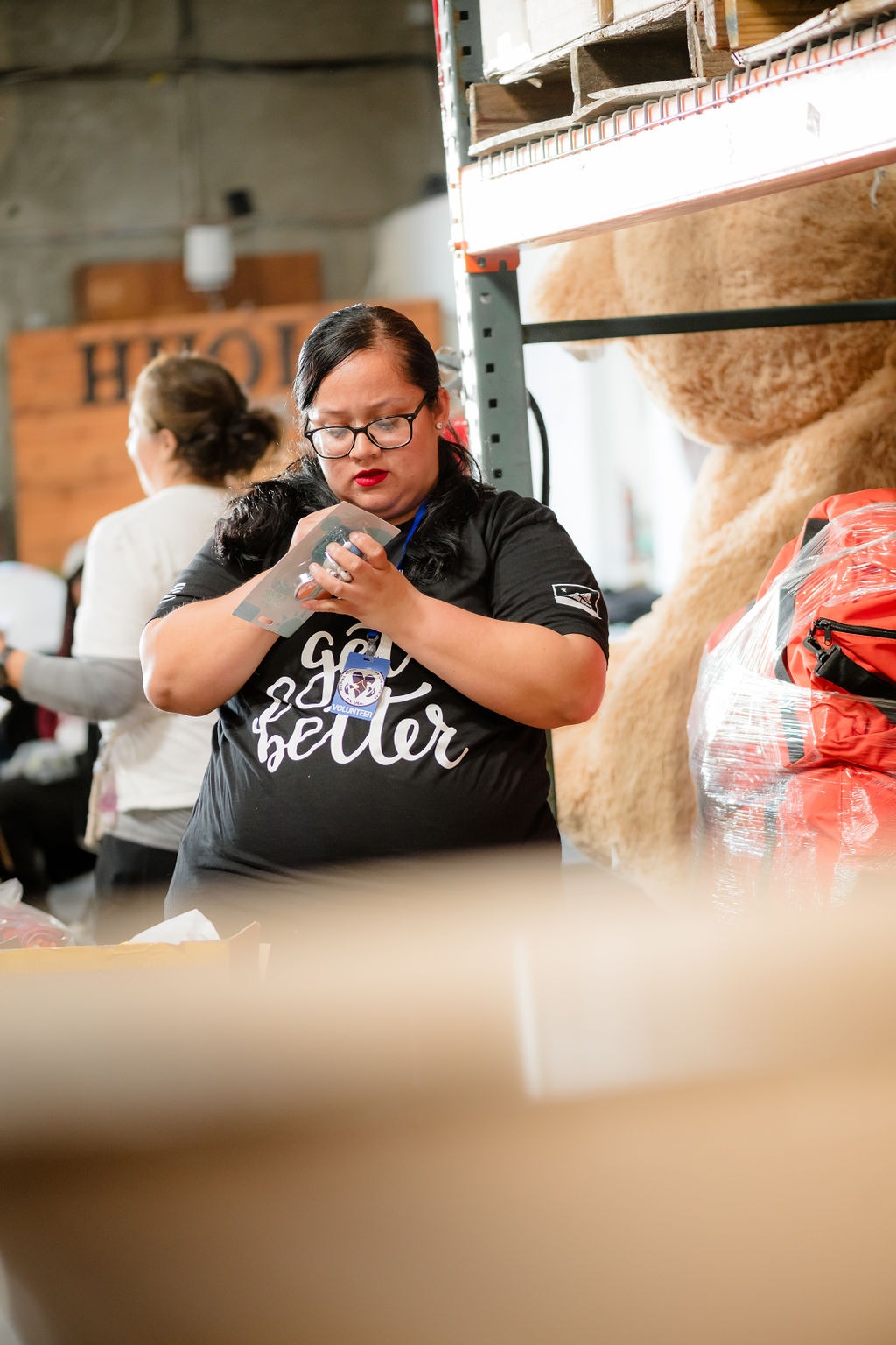 A volunteer inspects supplies in a warehouse, focusing on packaging details. A teddy bear and bags are visible in the background, highlighting the organization’s efforts.