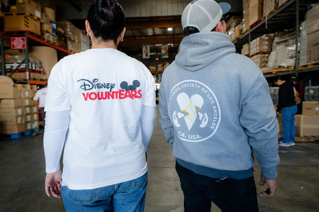 Two volunteers stand in a warehouse, wearing shirts promoting community service. One shirt features "Disney VOLUNTEARS," while the other represents the "Hand in Hand Society of Los Angeles."