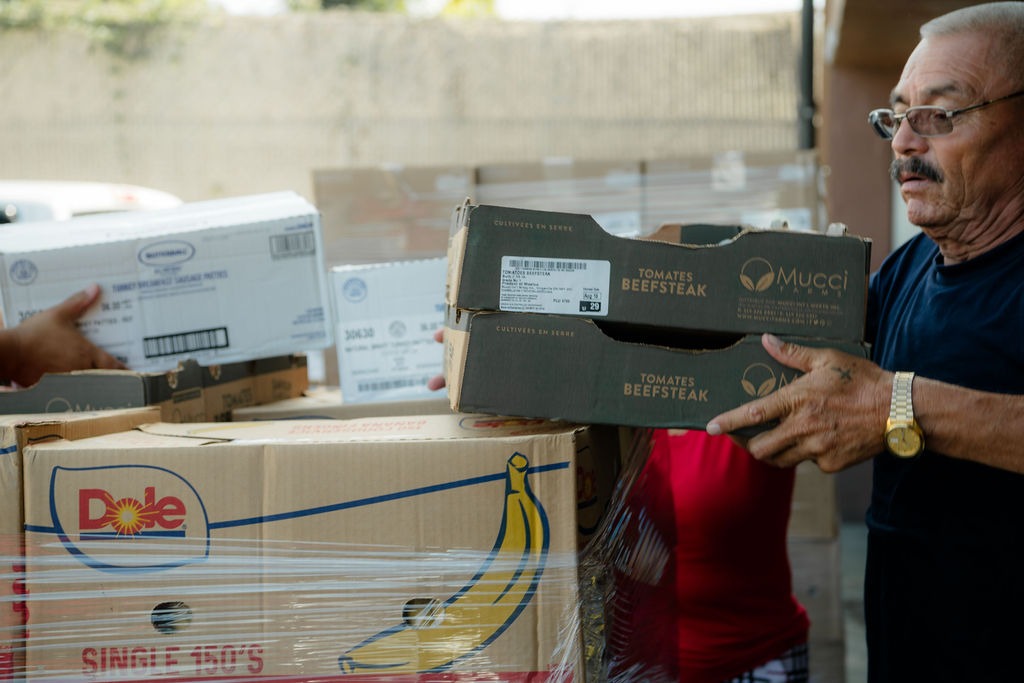 Two individuals transport boxes of food, including Dole banana cartons and tomato crates, highlighting community food distribution efforts.
