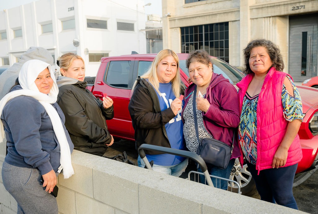 Five women stand together in a parking area, displaying friendly smiles. They wear casual clothing and show a sense of community, suggesting a social gathering or event.