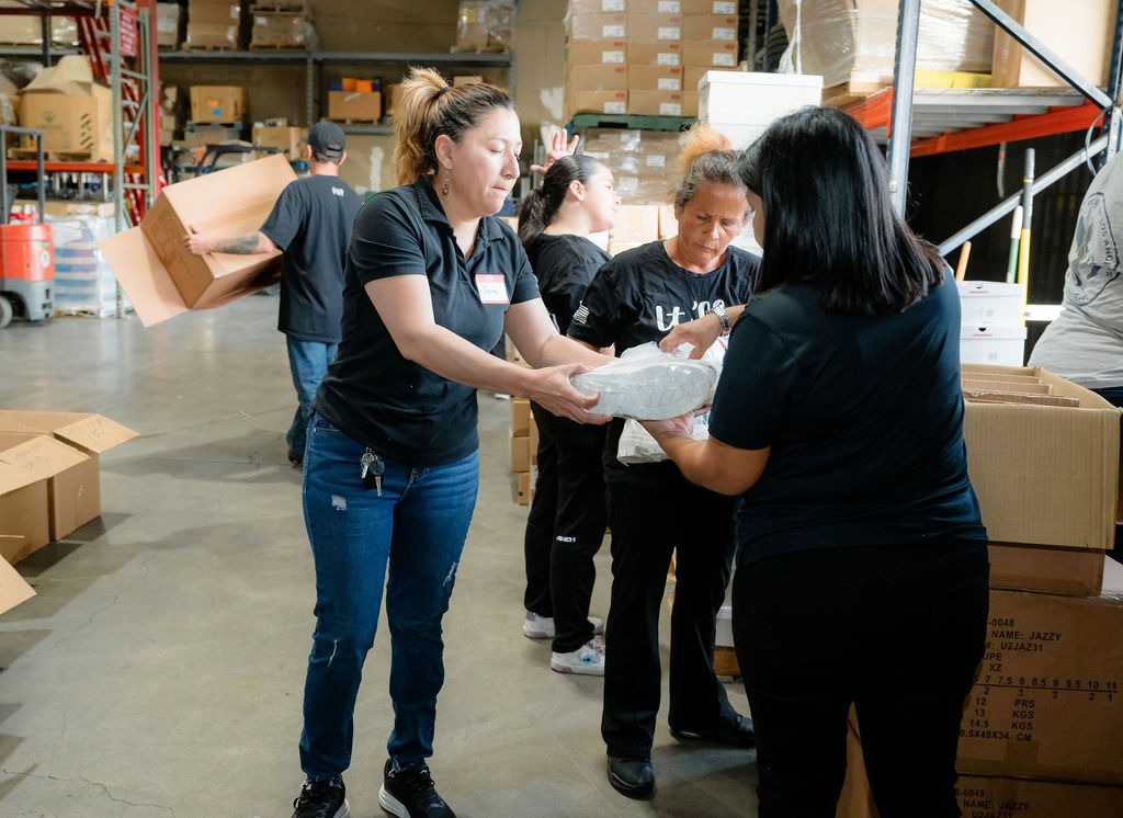 Volunteers in a warehouse distribute supplies, with two women passing a package. The scene highlights teamwork in community support efforts.