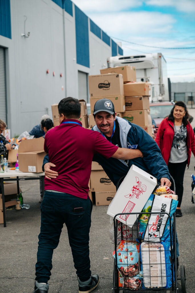 Two men embrace while handling food items outside a community food distribution event, with boxes stacked in the background.
