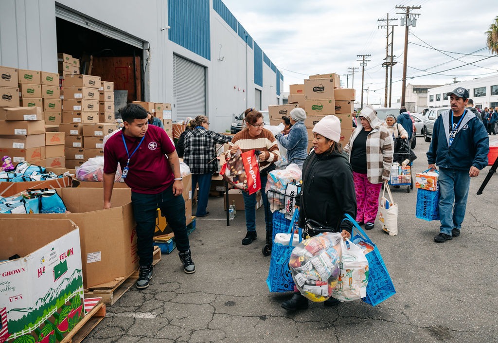 People gather outside a warehouse to collect groceries and supplies, reflecting community support during an outreach event.