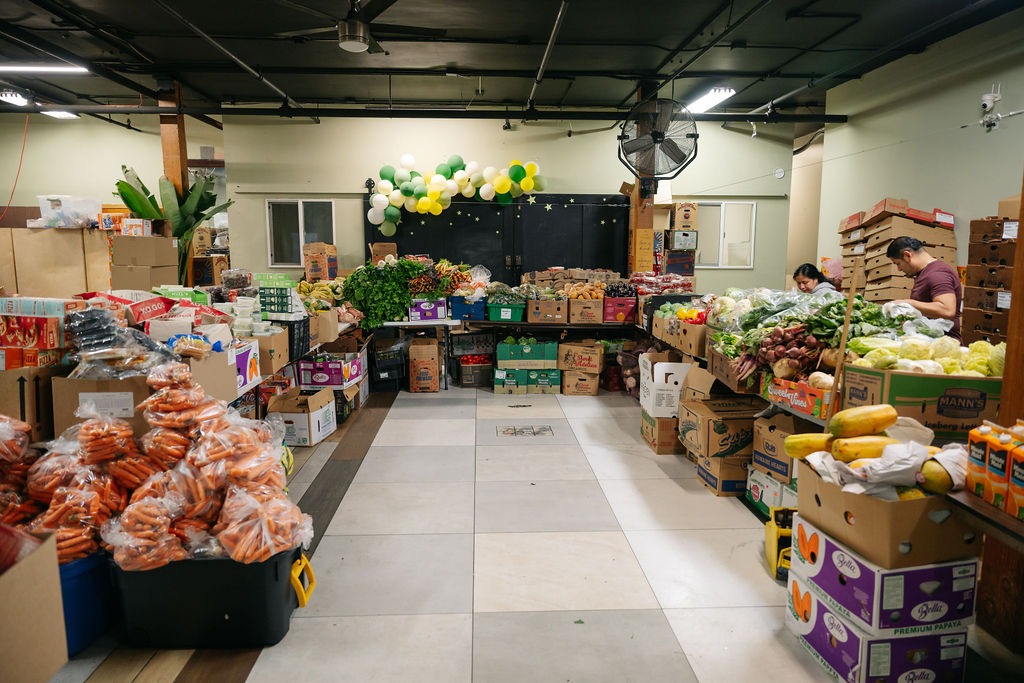 A vibrant community market filled with fresh produce, boxed goods, and customers. Colorful displays of fruits and vegetables create a lively shopping environment.